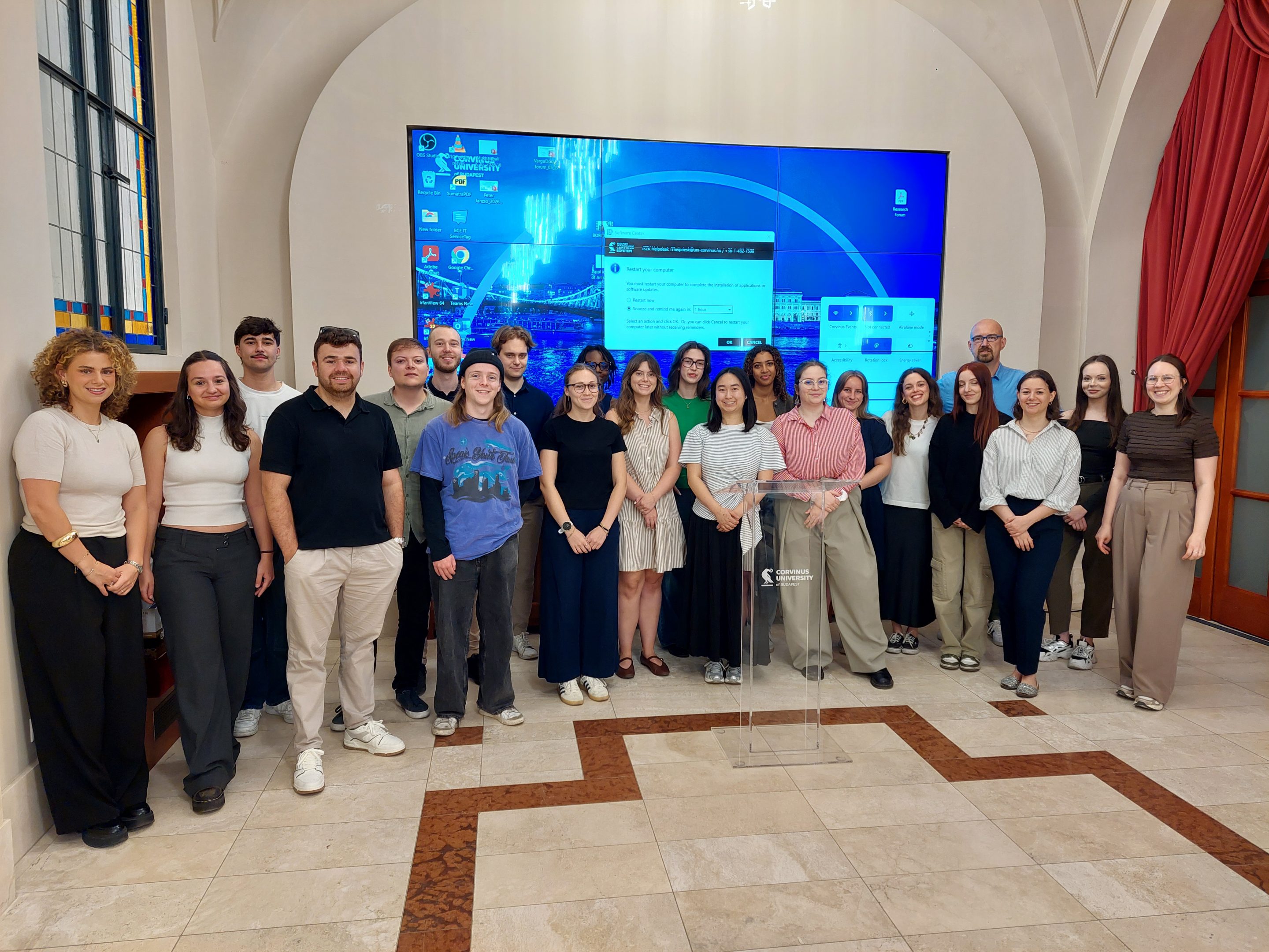 Worldcom Youth Meeting 2026 – equipo completo Group of students posing for a photo in front of a large mounted screen in a bright, spacious lobby or lecture hall.