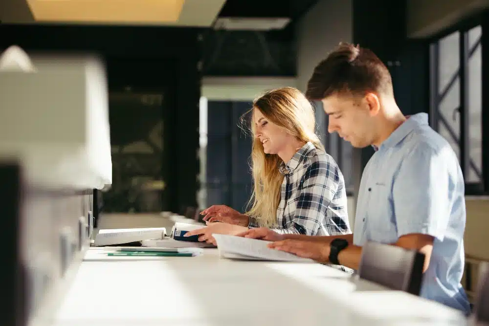 Two colleagues work at a bright office desk: a woman types on a keyboard while a man reviews papers nearby.