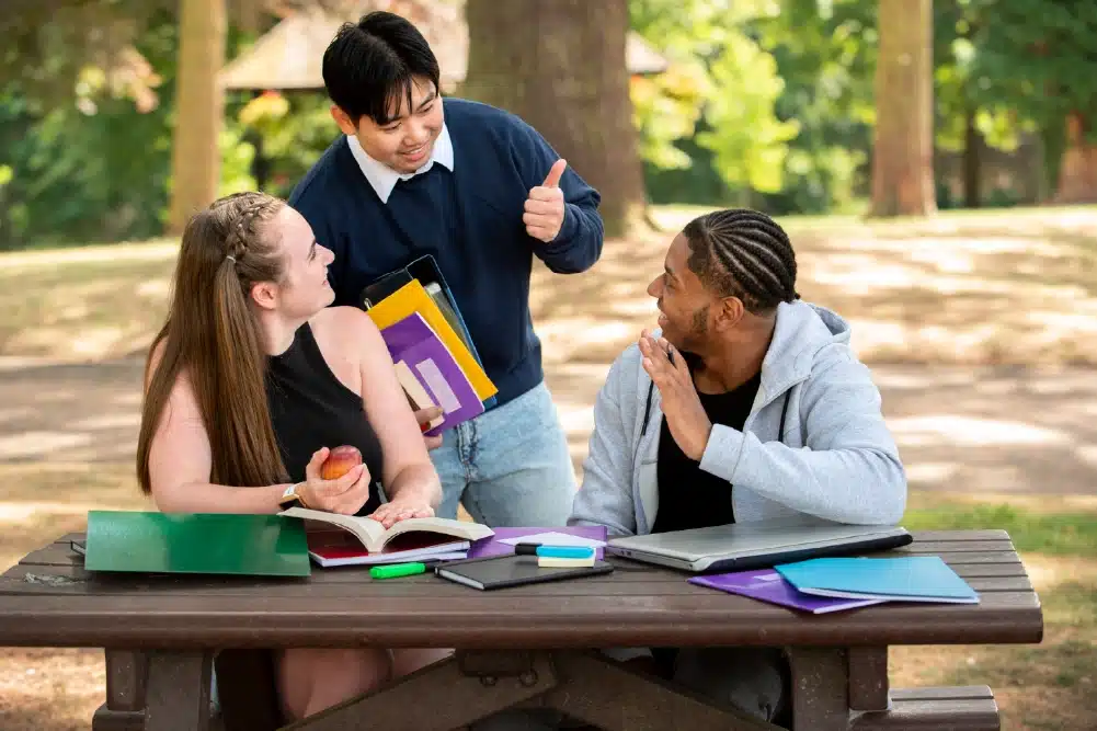 Three students studying at a park bench: two seated with books and notebooks, one standing and giving a thumbs up.