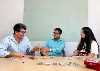 Three people sit around a table playing a card game, with colorful cards and tokens spread across the wooden table in a bright room.