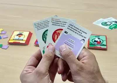 Hands hold three cards from a Spanish-language board game, with colorful tokens and game boxes on a wooden table in the background.
