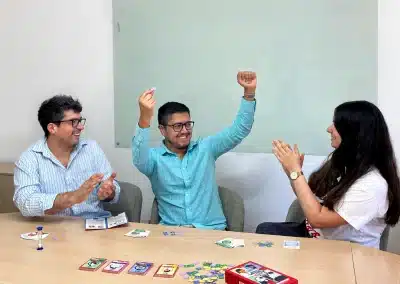 Three coworkers celebrate a win around a conference table while playing a board game, with cards and colorful pieces on the table.