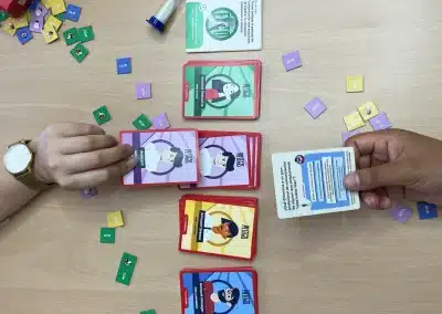 People play a colorful card game on a wooden table with role cards and small clip chips scattered around.