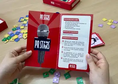 Hands hold a red game booklet with a microphone image and PR STARS logo; colorful square cards scattered on a wooden table behind it.